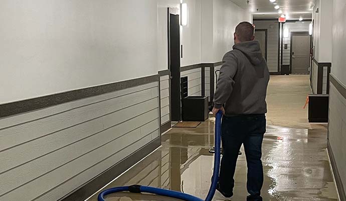 Expert using a blue hose to remove water from a flooded hallway with wet floors